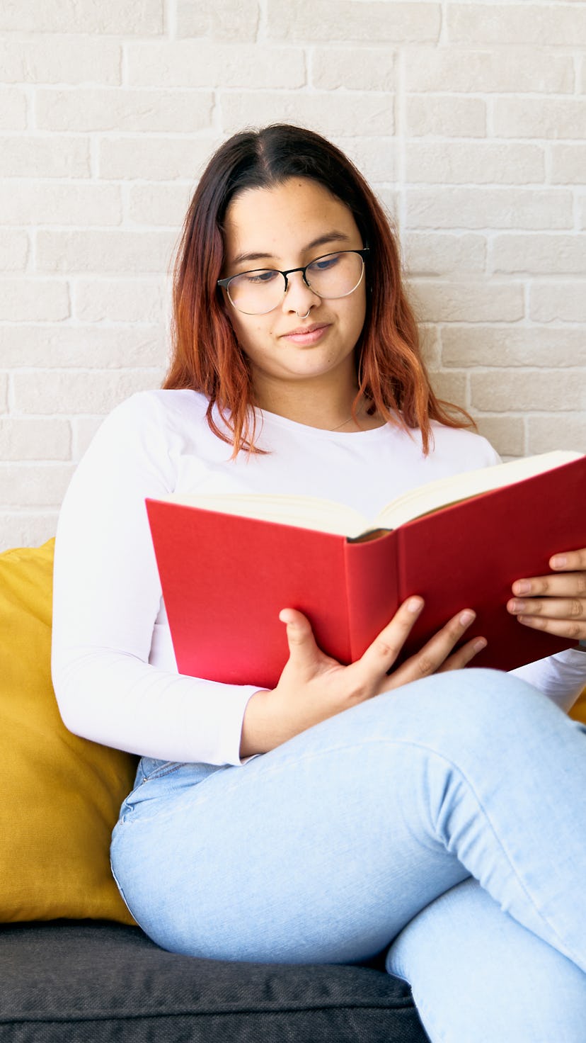 Beautiful teenage girl enjoying reading red book at home sitting on sofa with yellow cushions. She h...