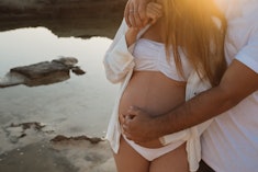 Cropped unrecognizable man embracing pregnant woman while standing on coast near calm sea in morning...