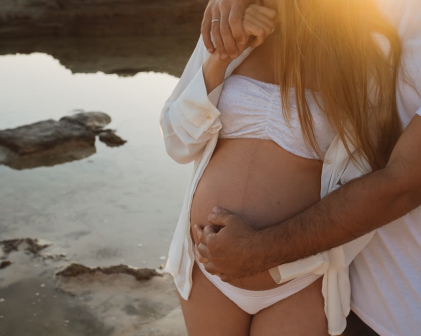 Cropped unrecognizable man embracing pregnant woman while standing on coast near calm sea in morning...