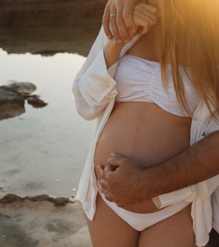 Cropped unrecognizable man embracing pregnant woman while standing on coast near calm sea in morning...