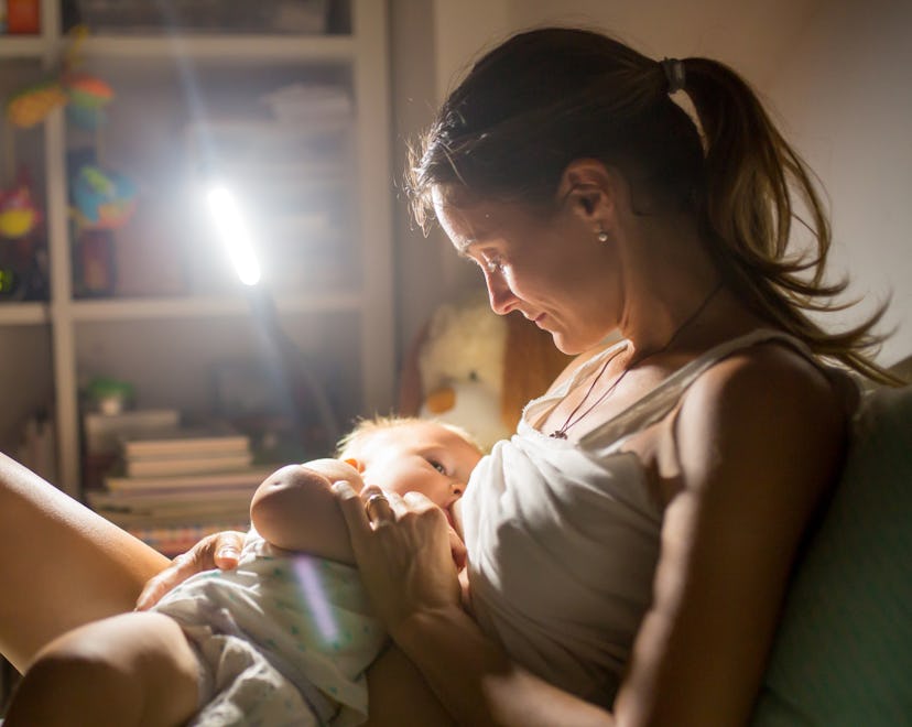 Young beautiful mother breastfeeding her newborn baby boy at night dim light. Mom breastfeeding infa...