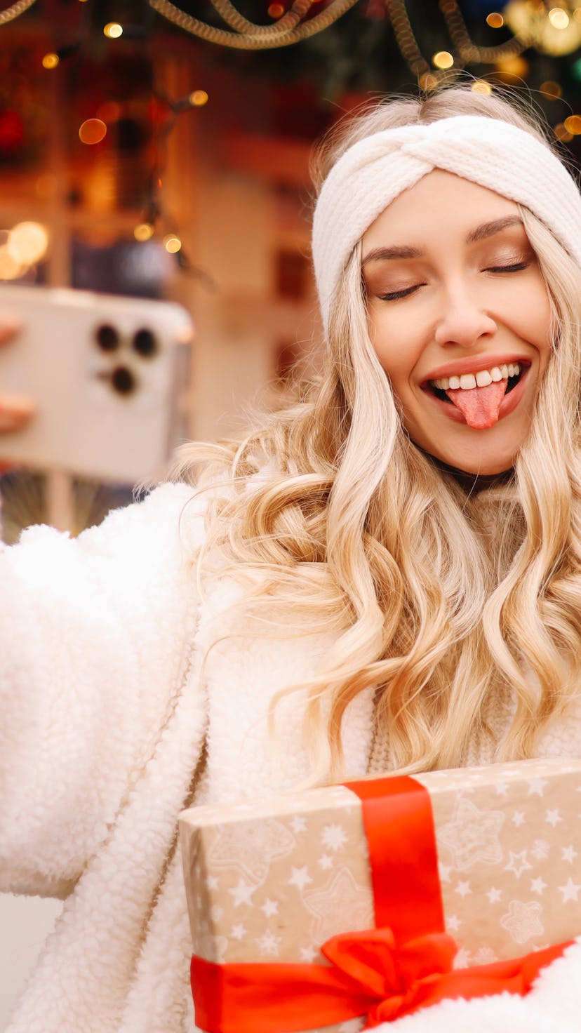 A happy blonde young woman in a hat and a warm wool coat holds a festive gift box in her hands, smil...