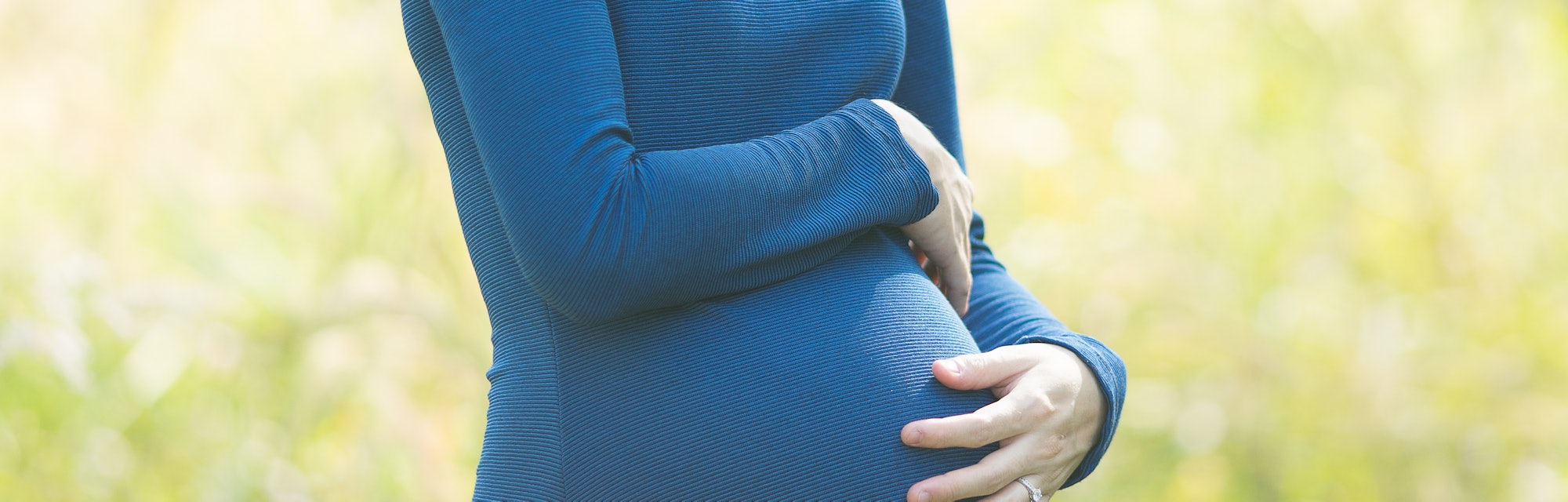 A pregnant woman with dark hair wearing a blue dress, smiling and cradling her belly, in an article ...