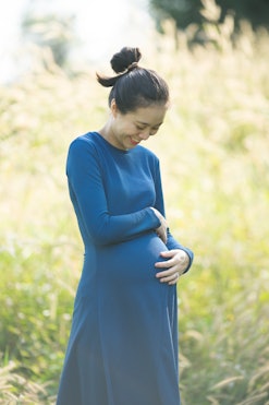 A pregnant woman with dark hair wearing a blue dress, smiling and cradling her belly, in an article ...