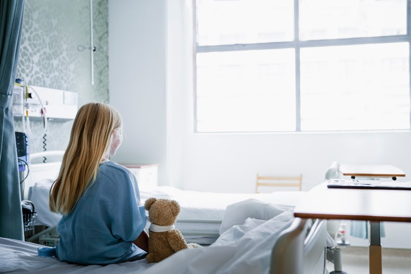 Little girl sitting on a hospital bed with her Teddy Bear by her side looking towards a window.
