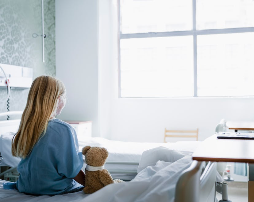 Little girl sitting on a hospital bed with her Teddy Bear by her side looking towards a window.