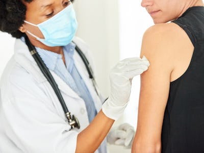 Female doctor with protective face mask and gloves disinfecting women patient's arm with cotton pad ...