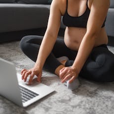 A pregnant woman works out by following an exercise video on her laptop in the modern, bright living...