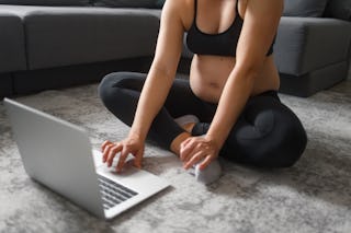 A pregnant woman works out by following an exercise video on her laptop in the modern, bright living...