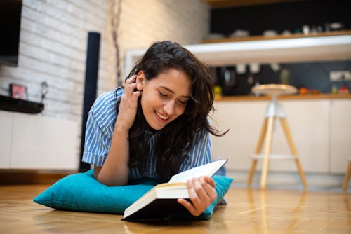 a beautiful young woman with dark brown hair lies on the floor, reading a book and smiling