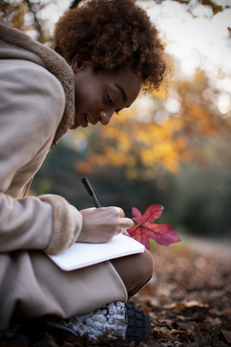 Young woman in coat with red autumn leaf writing in journal in park