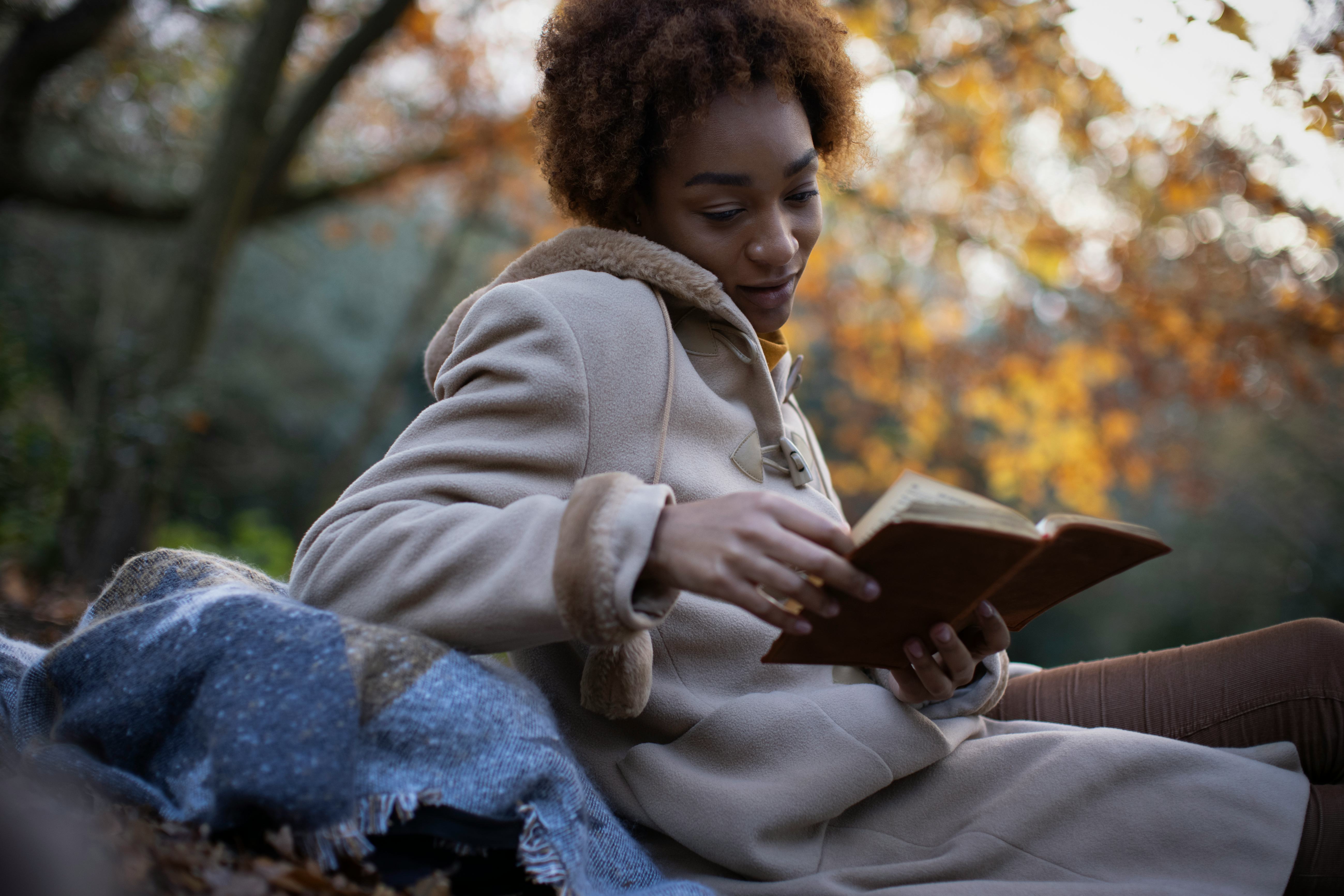 Young woman in brown coat reading book in autumn park