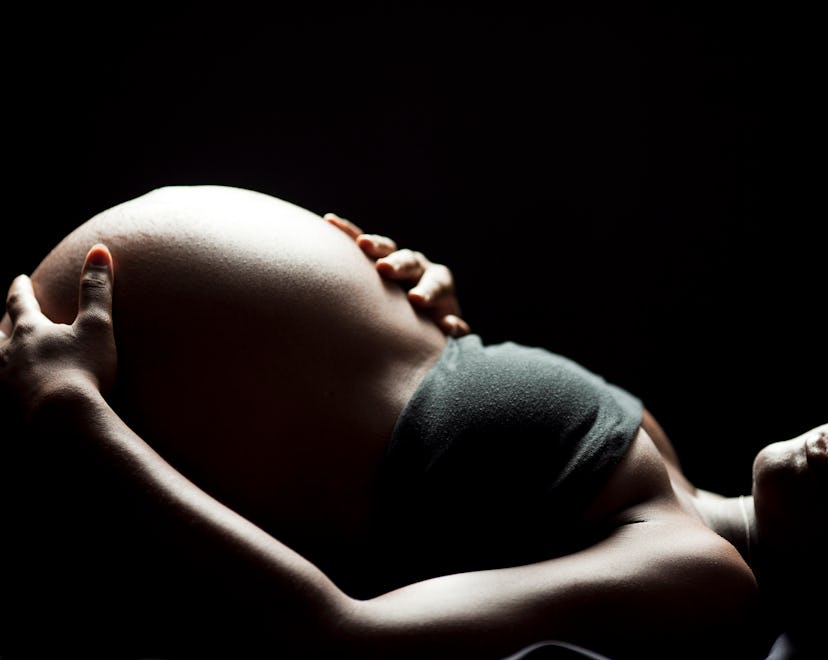 Pregnant woman with an exposed stomach lying down under heavy lighting in front of black background.