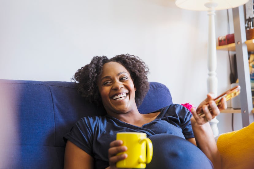 Laughing pregnant woman with tea and smart phone on sofa in an article about if baby boys are usuall...