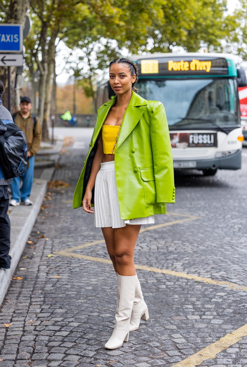 A guest wears green jacket, white pleated mini skirt, white boots, yellow cropped top outside Victor...