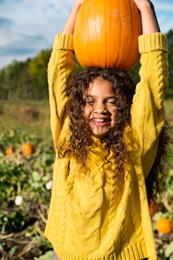A girl with long dark hair wearing a yellow sweater holds a pumpkin above her head and smiles at one...