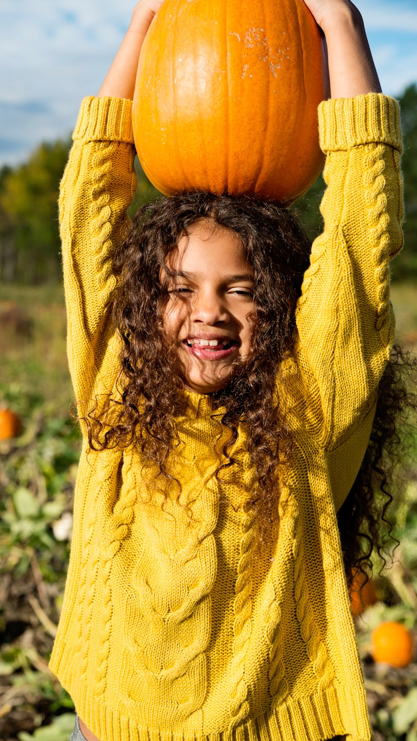 A girl with long dark hair wearing a yellow sweater holds a pumpkin above her head and smiles at one...