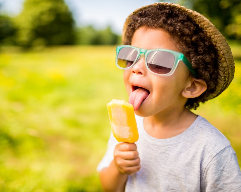 The tongue of a young boy with curly hair is touching an orange frozen treat.  He is wearing a gray ...