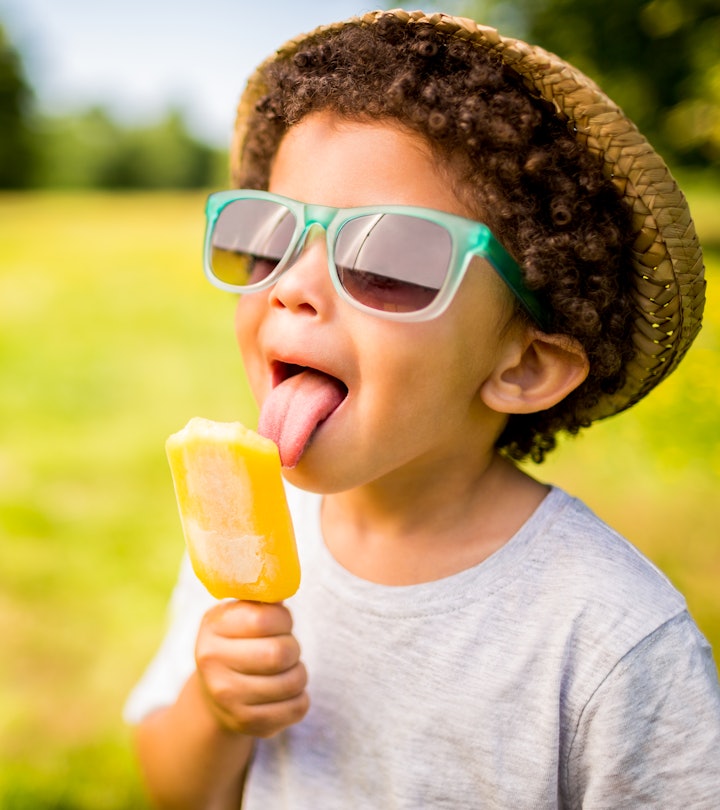 The tongue of a young boy with curly hair is touching an orange frozen treat.  He is wearing a gray ...