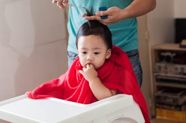 A dad standing behind his child in a high-chair, cutting their hair.