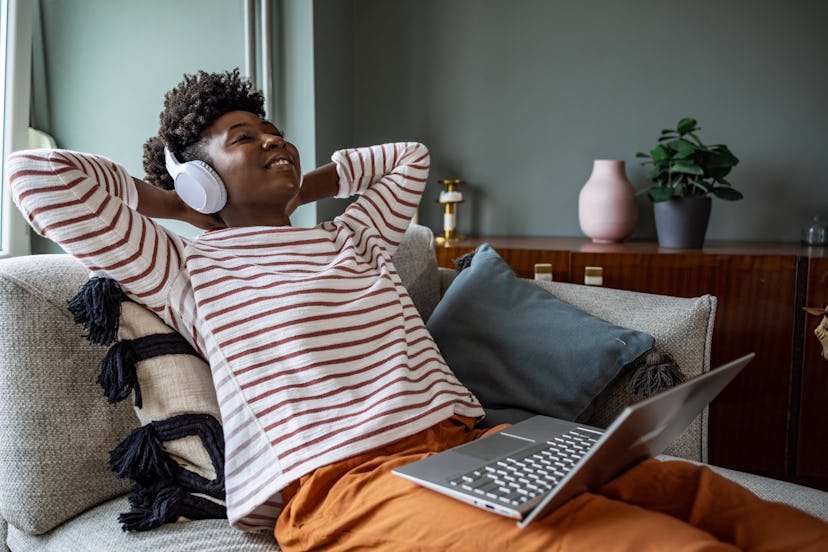 Young African American woman with wireless headphones relaxing on the couch and listening her favour...