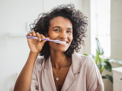 Young woman in pajamas looking in mirror and brush teeth with toothbrush in home bathroom.