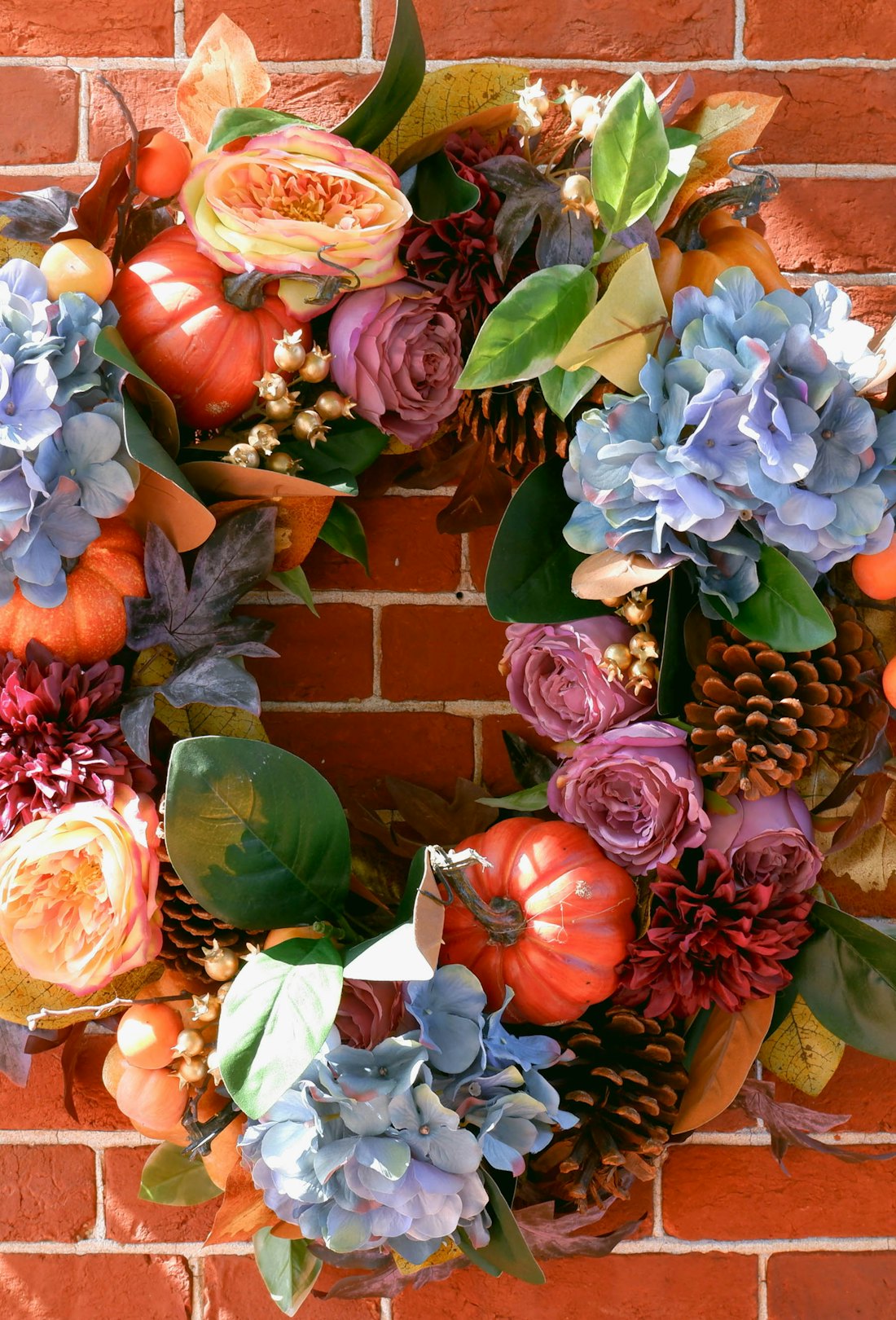 Close-up of a fall wreath on brick wall with blue and purple flowers, pumpkins, and greenery.