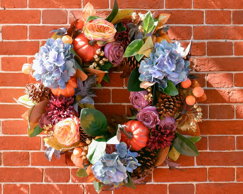 Close-up of a fall wreath on brick wall with blue and purple flowers, pumpkins, and greenery.