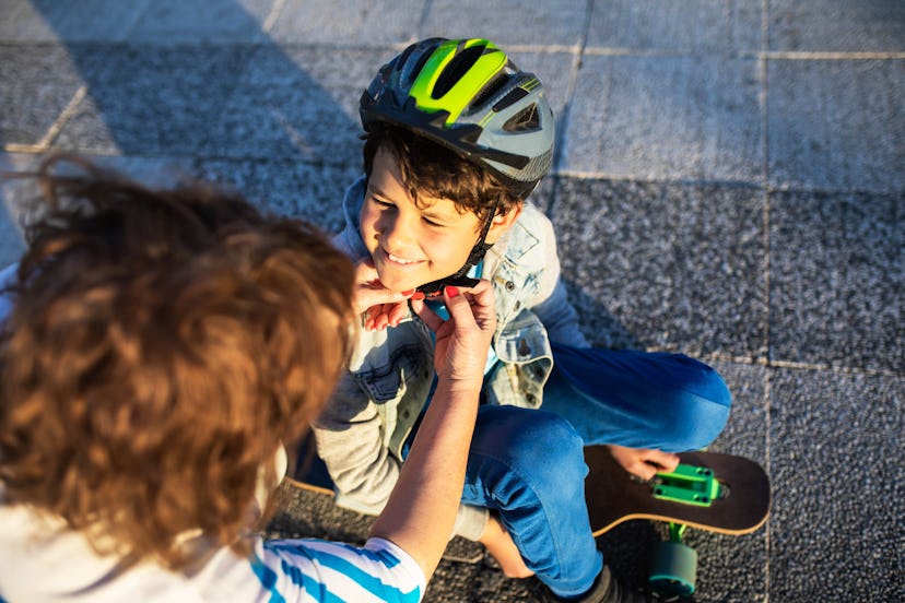 A cute boy is smiling on a longboard while his nanny is fastening his helmet in a piece about how to...