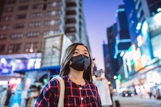 Lovely girl with protective face mask looking up to sky while walking in downtown city street at nig...