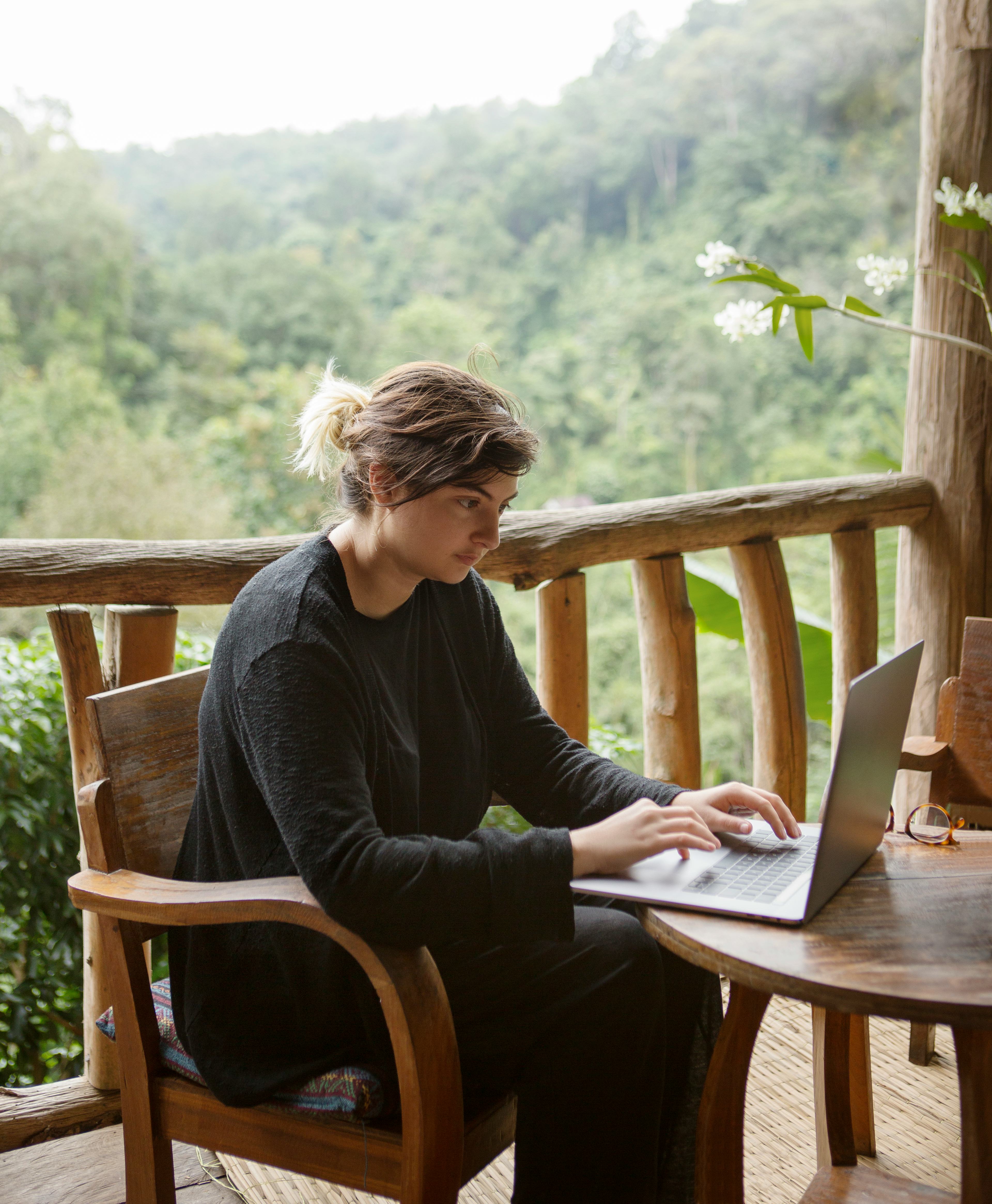 A young woman works on her laptop at a wooden table overlooking the misty mountain jungles of Doi Su...