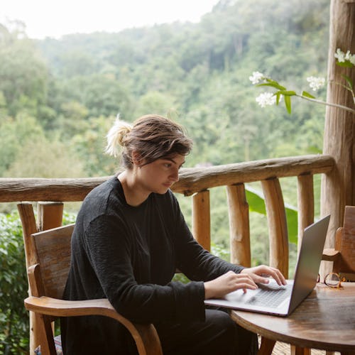 A young woman works on her laptop at a wooden table overlooking the misty mountain jungles of Doi Su...
