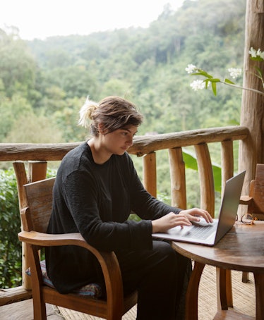 A young woman works on her laptop at a wooden table overlooking the misty mountain jungles of Doi Su...