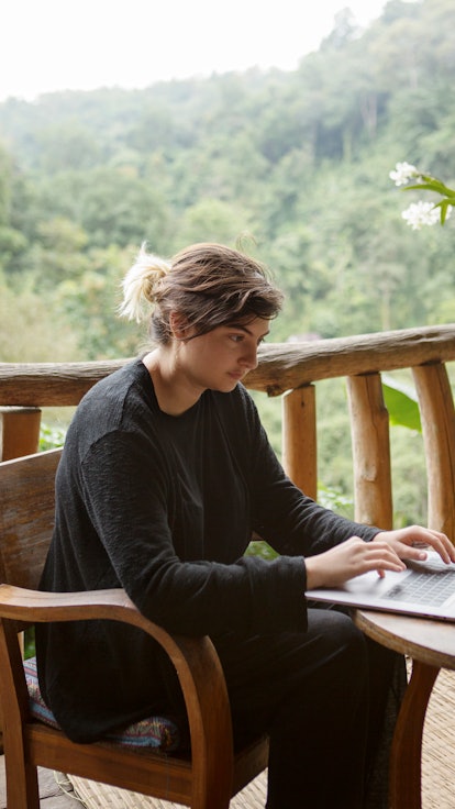 A young woman works on her laptop at a wooden table overlooking the misty mountain jungles of Doi Su...