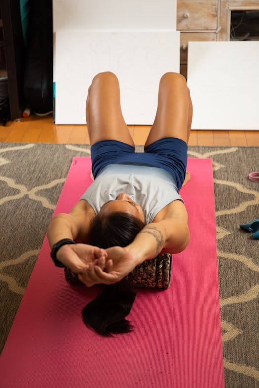 A fit tanned sporty woman rolling her shoulders and back over a foam roller a fitness mat in her liv...
