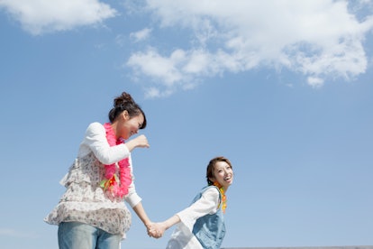 Two young women in jeans and colorful necklaces laugh and dance with each other against a blue sky w...
