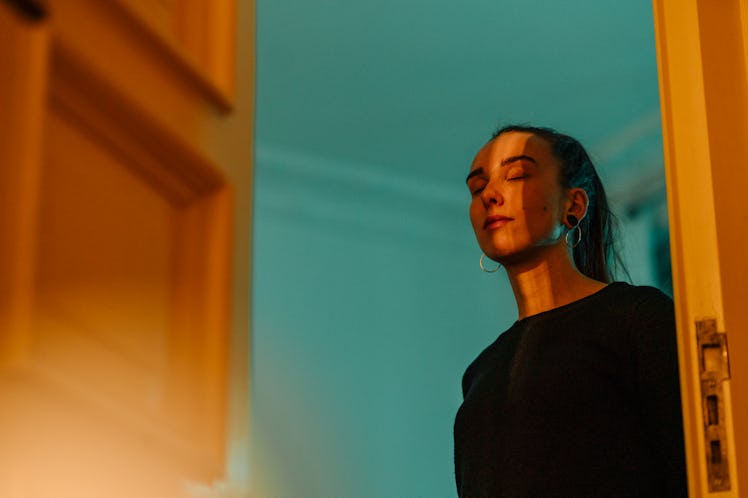 A young woman with her hair in a ponytail stands with her eyes closed in an open doorway, the room b...