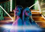 Two young blonde women wearing sunglasses sit leaning back-to-back on a staircase lit by neon lights...