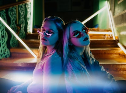 Two young blonde women wearing sunglasses sit leaning back-to-back on a staircase lit by neon lights...