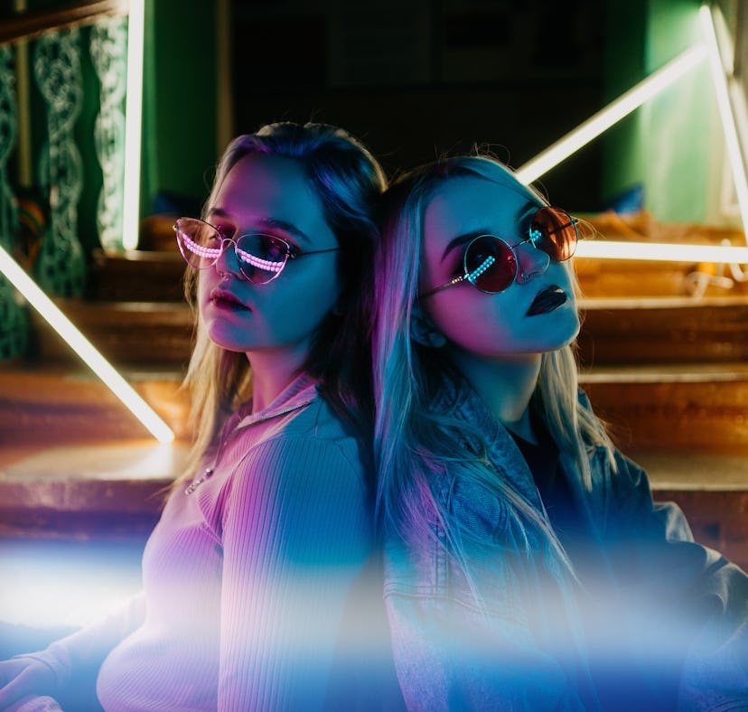 Two young blonde women wearing sunglasses sit leaning back-to-back on a staircase lit by neon lights...