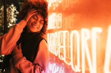 A smiling young woman with curly hair leans against an orange neon sign so the light reflects up on ...