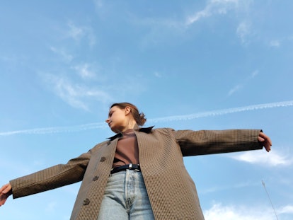 A young woman in a long coat and jeans is seen from below, posing with her arms spread wide as she l...