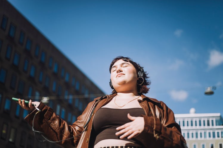 A young woman with short dark hair closes her eyes and dances to music in a city landscape against a...