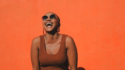 A young woman with a shaved head laughs heartily in front of an orange background as she considers t...