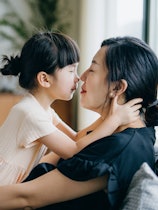 Side profile of lovely little Asian girl embracing and kissing her mother while relaxing at home. Bo...