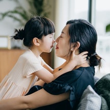 Side profile of lovely little Asian girl embracing and kissing her mother while relaxing at home. Bo...