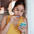 Mixed race young woman, brushing teeth and checking phone outside bathroom