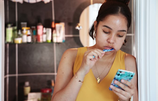 Mixed race young woman, brushing teeth and checking phone outside bathroom