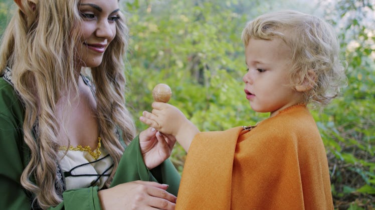 Fairy elf cosplay woman treats little baby boy hobbit with candy in green forest. Halloween concept,...