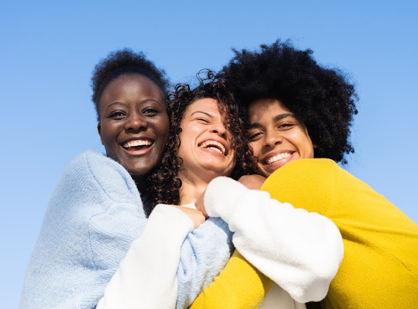 Three cheerful multi-ethnic young women embracing on blue background and two of them looking at the ...